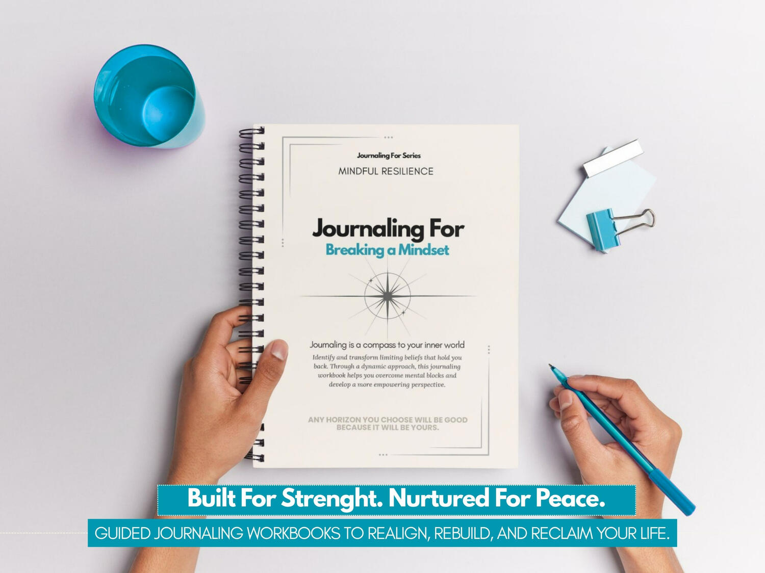 Journaling For – Breaking a Mindset’ spiral-bound workbook held by a person on a clean white desk with teal stationery elements.